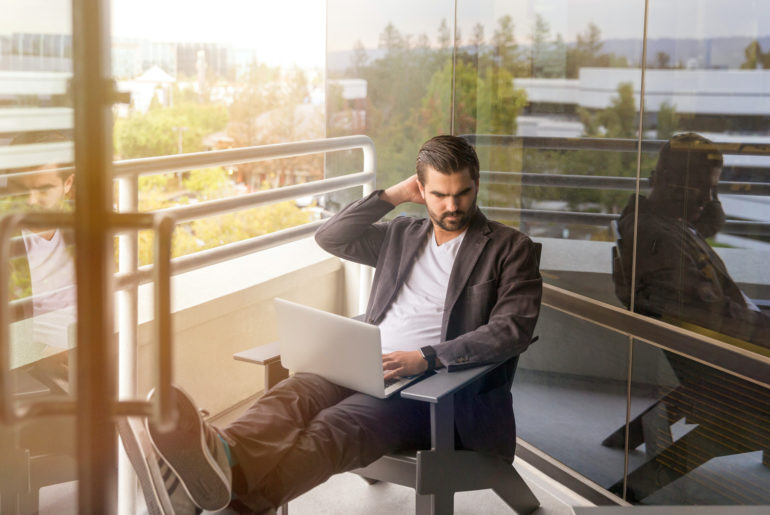 A man using a laptop computer for remote work while sitting on a bench.