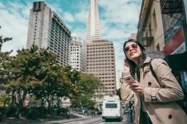 A woman in a trench coat is looking at her phone while standing on a street in San Francisco, seemingly unaware of the bustling movement around her.
