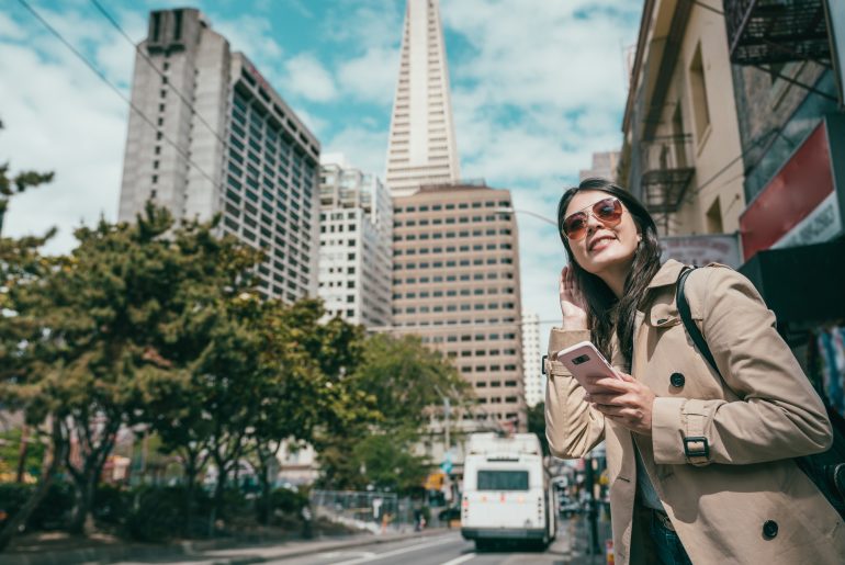A woman in a trench coat is looking at her phone while standing on a street in San Francisco, seemingly unaware of the bustling movement around her.