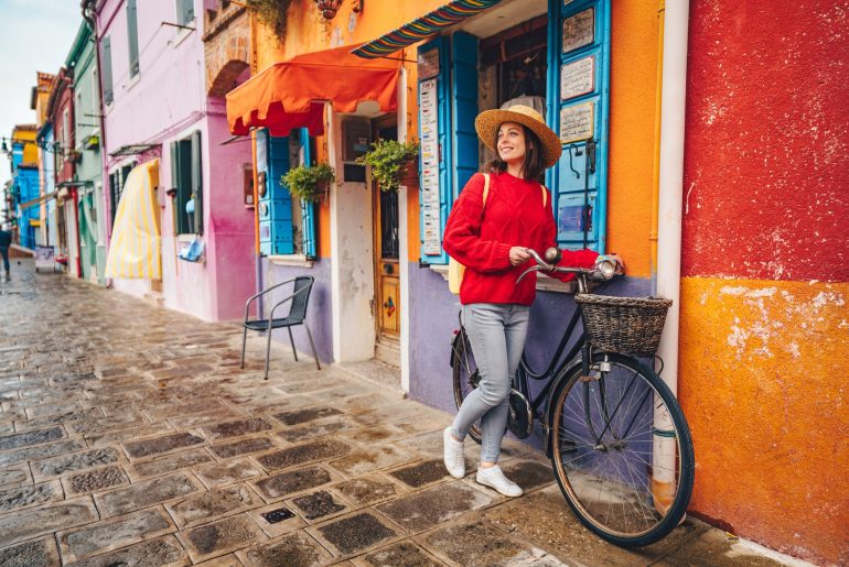 A woman is leaning on a bicycle in front of colorful houses in Burano, Italy during relocation.