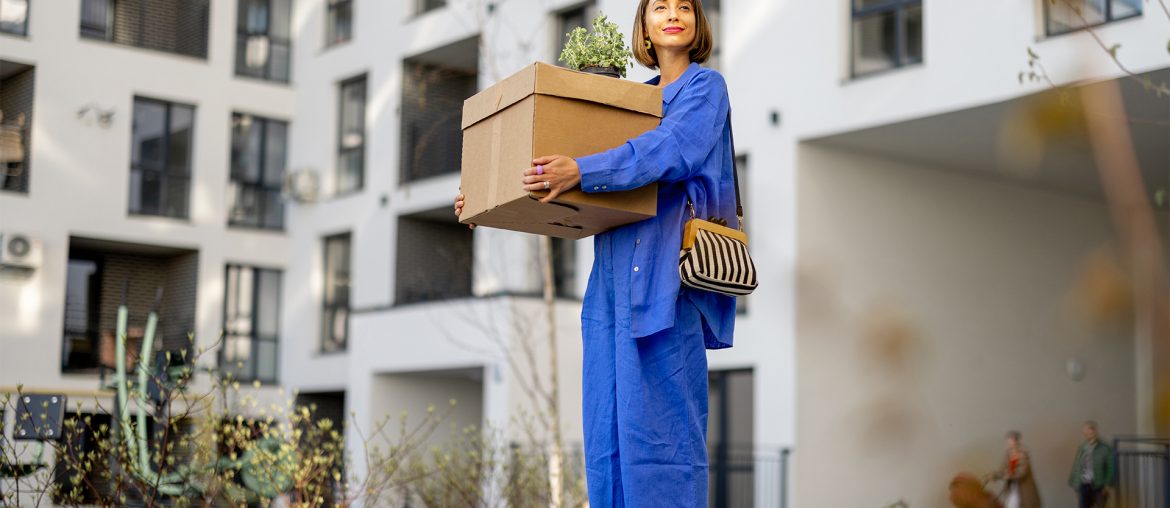 A woman in a blue jumpsuit holding a box and purse, standing in an apartment complex courtyard, facing the camera.