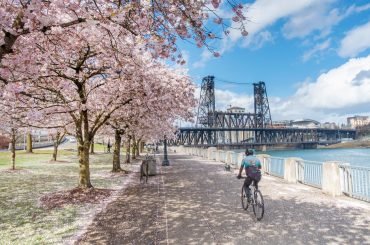 Eco-Relocation: 7 of the Greenest Cities in the U.S. A person biking under cherry blossom trees along a river with a steel bridge in the background on a sunny day after a recent relocation.