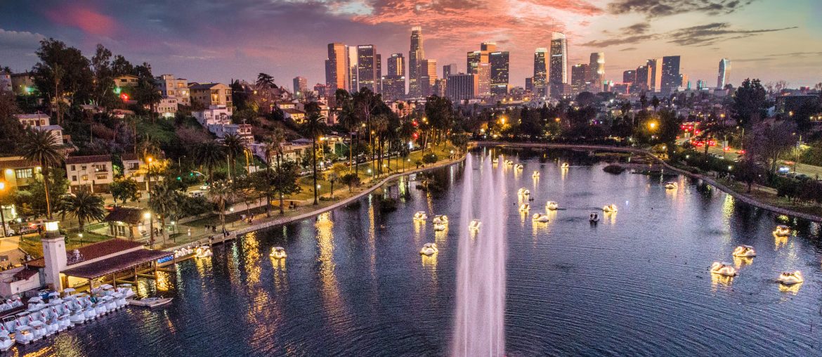 Aerial view of a park lake with lit-up fountains and swan pedal boats, set against the Los Angeles city skyline at sunset.