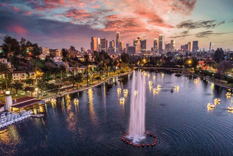 Aerial view of a park lake with lit-up fountains and swan pedal boats, set against the Los Angeles city skyline at sunset.