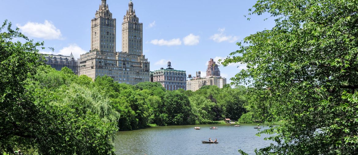 A picturesque view of Central Park from the Upper West Side featuring a pond, people, and boats, framed by greenery, with towering twin skyscrapers in the background.