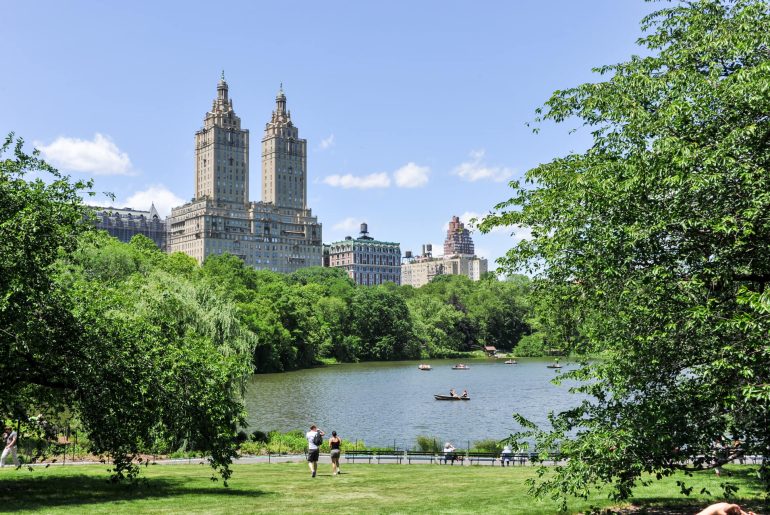A picturesque view of Central Park from the Upper West Side featuring a pond, people, and boats, framed by greenery, with towering twin skyscrapers in the background.