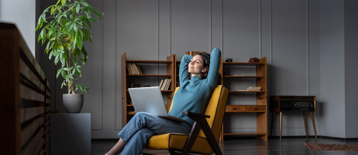 A person sits in a chair with their hands behind their head, looking relaxed. A laptop rests on their lap, hinting at remote work. The background features shelves and a potted plant in a modern room, creating an atmosphere that promotes mental health and well-being.