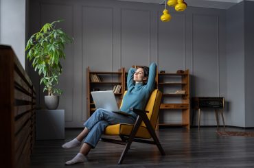 A person sits in a chair with their hands behind their head, looking relaxed. A laptop rests on their lap, hinting at remote work. The background features shelves and a potted plant in a modern room, creating an atmosphere that promotes mental health and well-being.