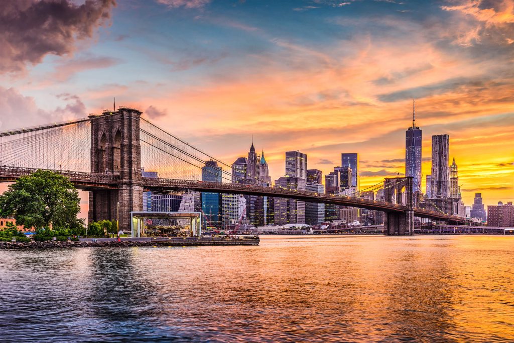 Brooklyn Bridge in New York City with the Manhattan skyline in the background at sunset, a perfect backdrop for a digital nomad. The sky is colorful with shades of orange, pink, and purple reflecting on the water below.