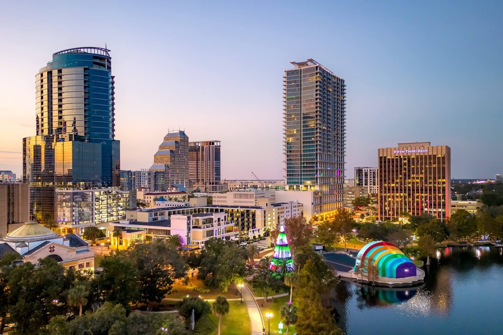 Aerial view of downtown Orlando at dusk, featuring high-rise buildings, a lit amphitheater with rainbow colors, and a decorated park with greenery and water; an inviting scene for any digital nomad seeking a vibrant urban environment to work and unwind.