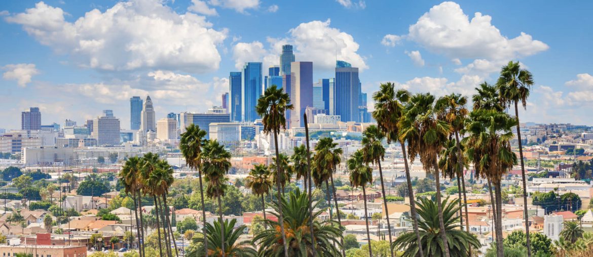 Downtown Los Angeles skyline with tall buildings and palm trees in the foreground, under a blue sky with scattered clouds.