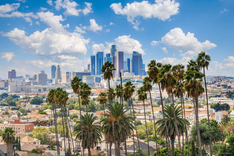 Downtown Los Angeles skyline with tall buildings and palm trees in the foreground, under a blue sky with scattered clouds.