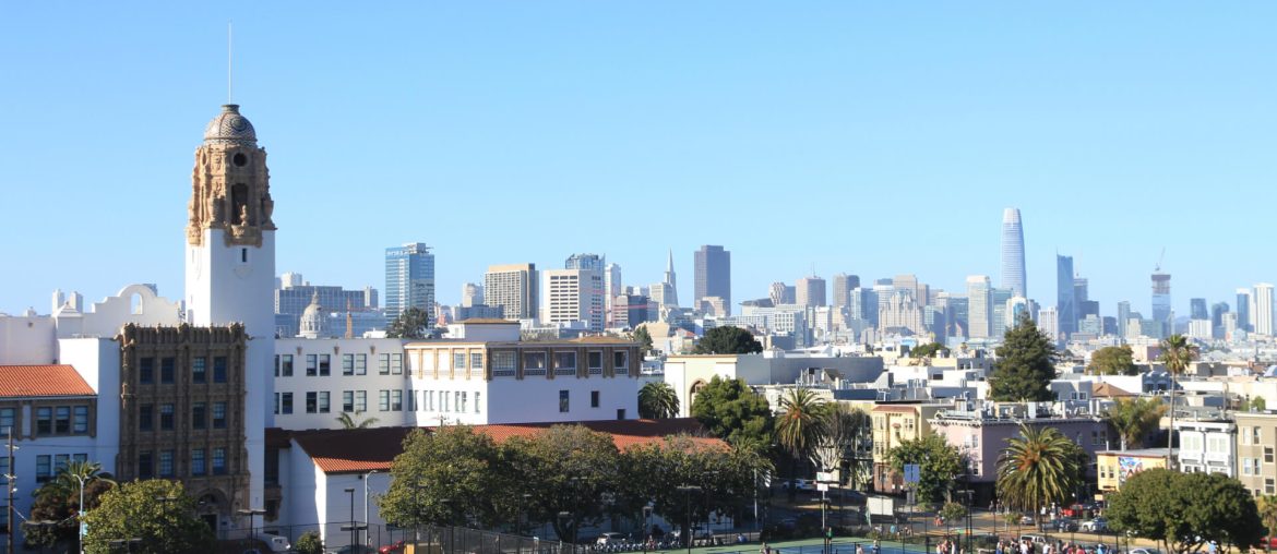 A city park with many people sitting on the grass, historic building on the left, and skyline of modern high-rise buildings in the background under a clear blue sky.