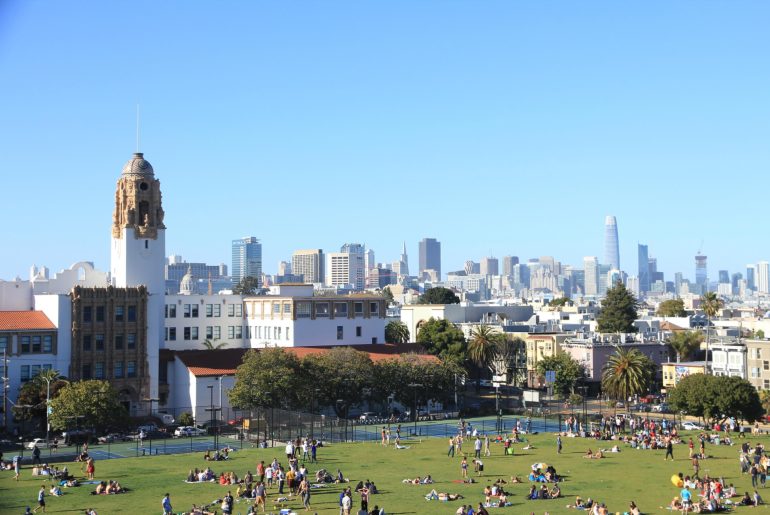 A city park with many people sitting on the grass, historic building on the left, and skyline of modern high-rise buildings in the background under a clear blue sky.