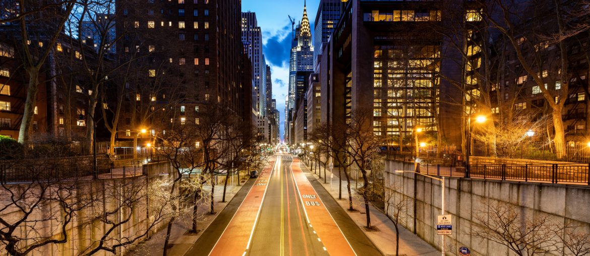 A city street in Murray Hill, Manhattan is flanked by tall buildings and illuminated at dusk. The road is mostly empty, with streetlights casting a warm glow. The Chrysler Building is visible in the distant background.