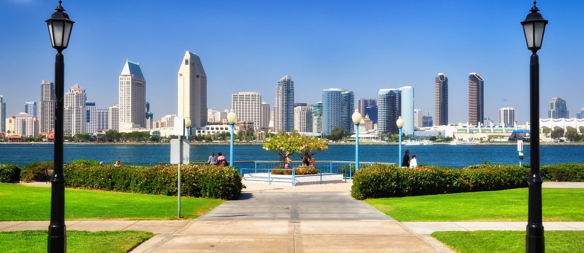 A view of a city skyline featuring modern high-rise buildings across a body of water, framed by two street lamps and green lawns on either side of a paved pathway.