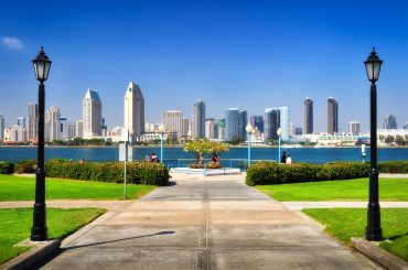 Moving to San Diego: The Essential Relocation Guide A view of a city skyline featuring modern high-rise buildings across a body of water, framed by two street lamps and green lawns on either side of a paved pathway.