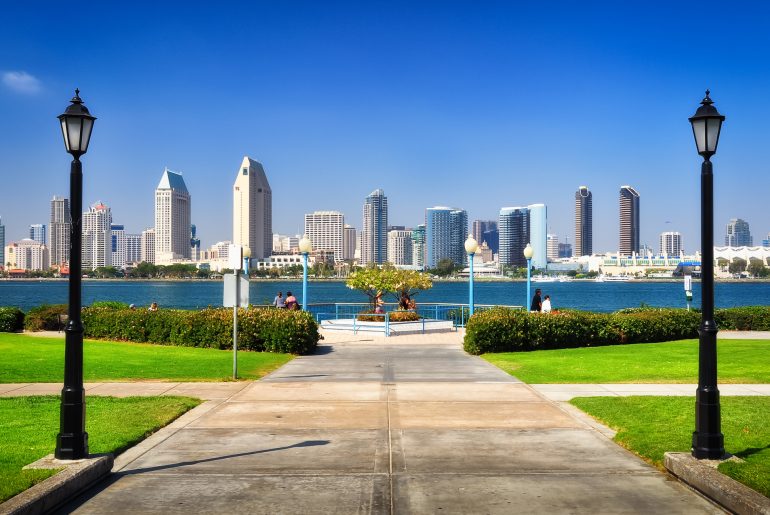 A view of a city skyline featuring modern high-rise buildings across a body of water, framed by two street lamps and green lawns on either side of a paved pathway.