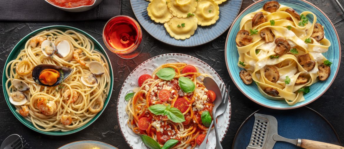 An assortment of pasta dishes, including ravioli, spaghetti with seafood, fettuccine with mushrooms, and tortellini, served with sauces and garnished with basil. Multiple utensils and glasses of drinks nearby. Experience this culinary delight on our exclusive food tour.