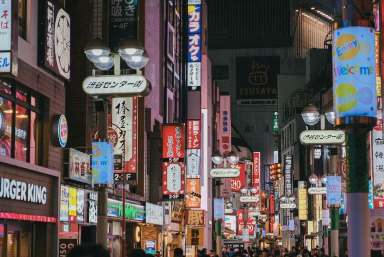 Colorful neon-lit street in Tokyo's busy Shibuya district, crowded with people and shops.