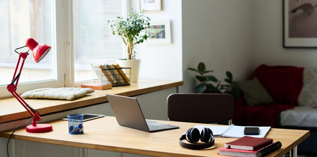 Home office desk with a laptop, headphones, and a red desk lamp next to a window in a furnished apartment