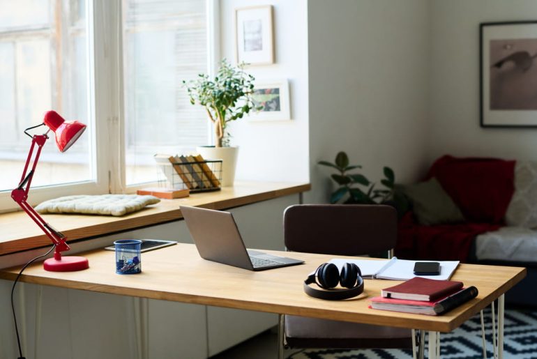 Home office desk with a laptop, headphones, and a red desk lamp next to a window in a furnished apartment